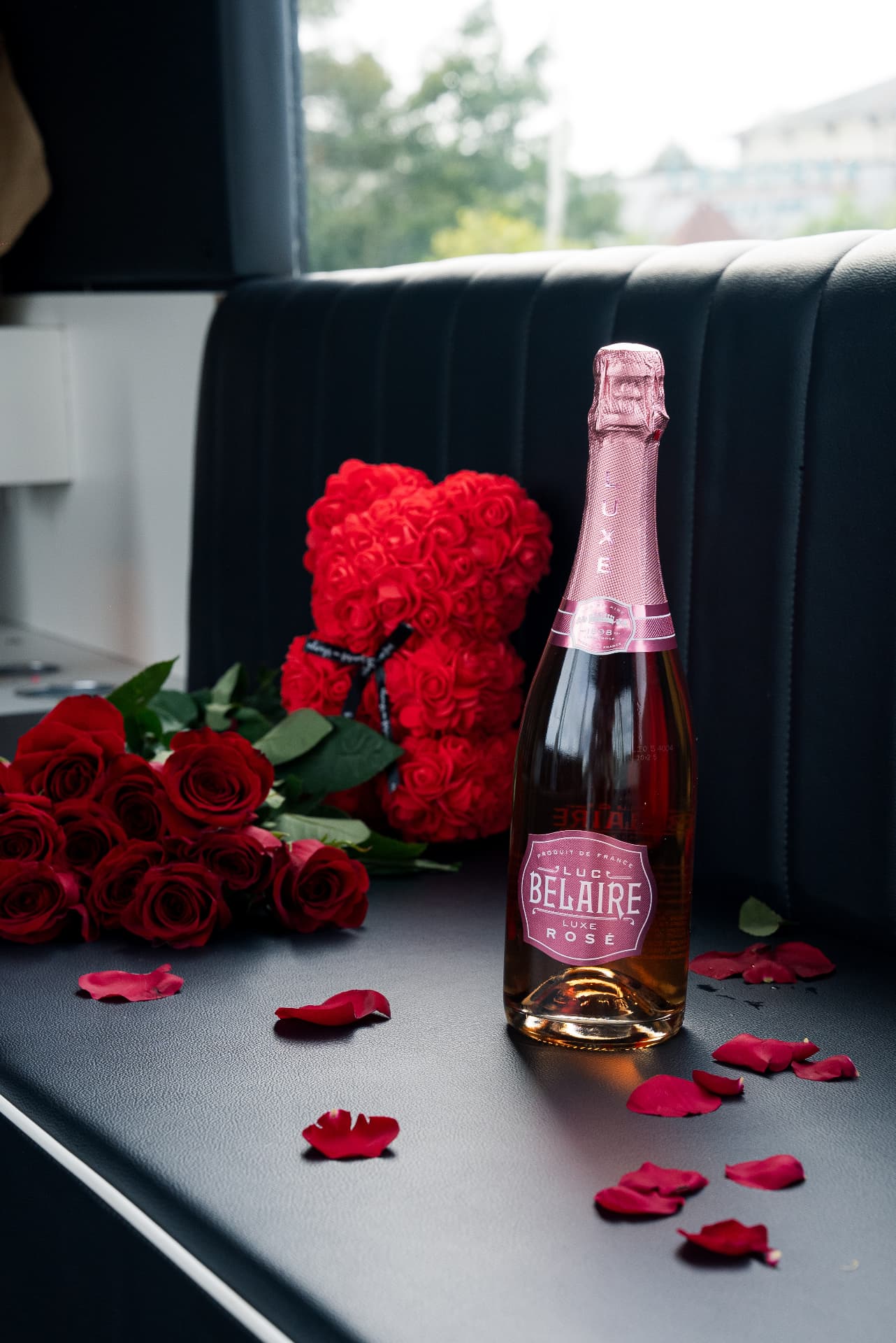 Bottle of Luc Belaire Luxe Rosé beside a rose bear and rose petals on the black leather bench
